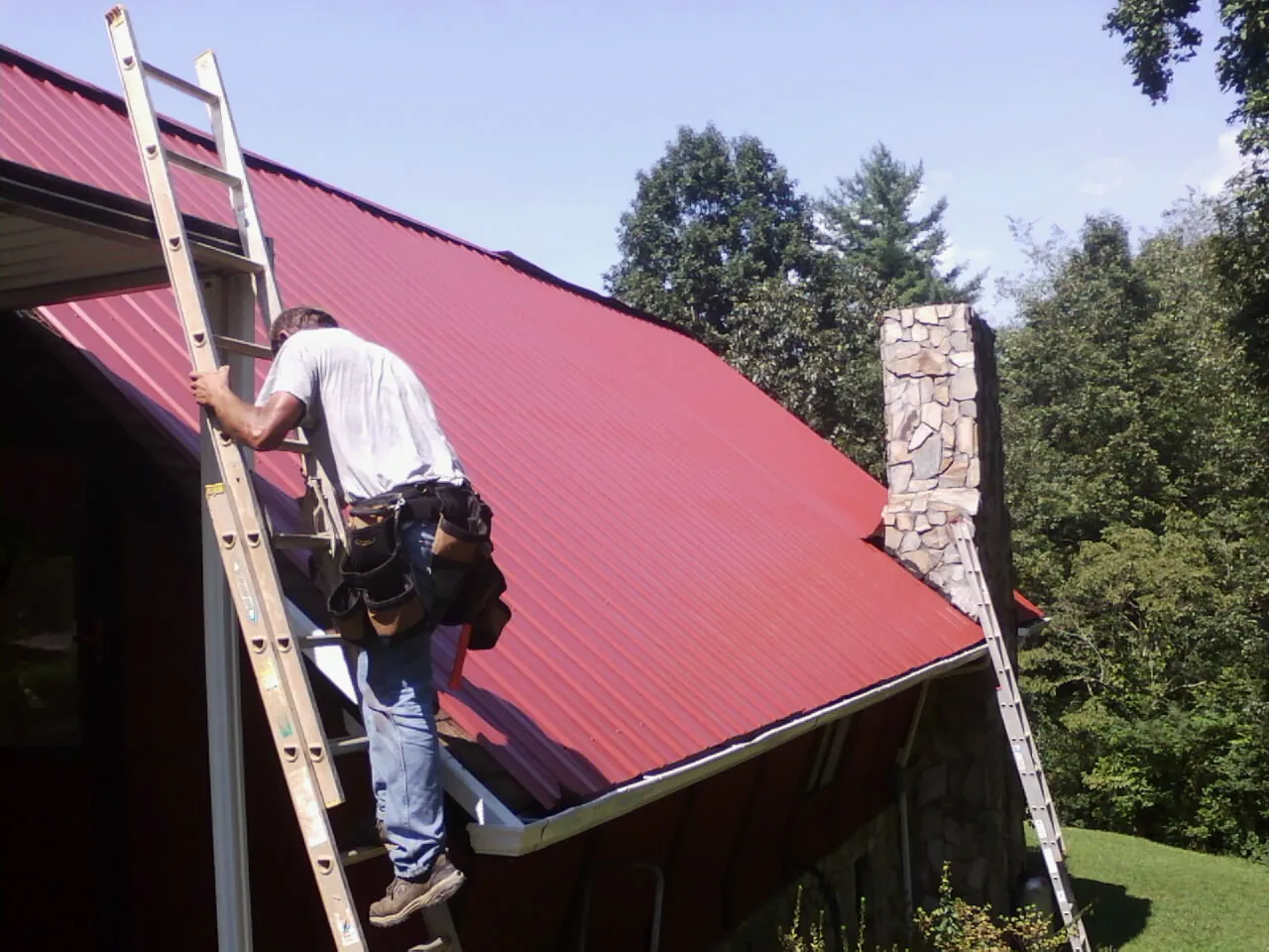 Roofer on a ladder beside a red metal roof and stone chimney — Soffit & Fascia Repair by Roofing Brothers in Pittsville